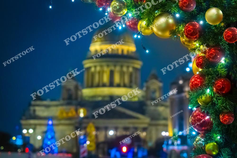 На переднем плане видны яркие рождественские украшения с красными и золотыми шарами, а также гирлянды. На заднем плане возвышается Исаакиевский собор, подсвеченный теплым светом. Атмосфера новогоднего Петербурга подчеркнута праздничной иллюминацией и праздничным декором. Эта сцена передает ощущение уюта и волшебства в зимнем городе.